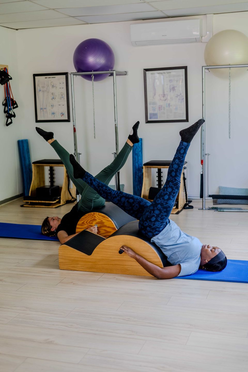 Two instructors doing a leg exercise on a spine corrector on blue mats in a Pilates class at Pilates Dynamics
