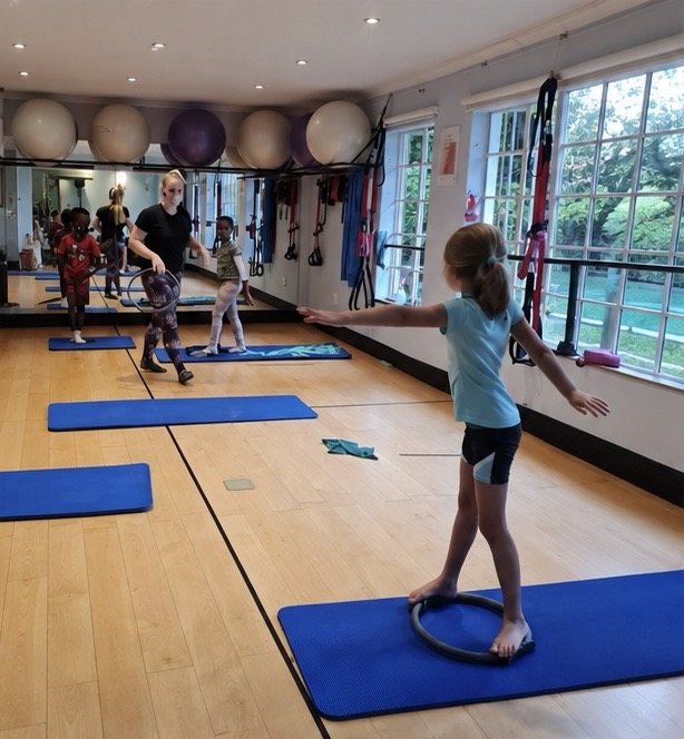 Children balancing on Pilates rings on their mats, an instructor guiding the class